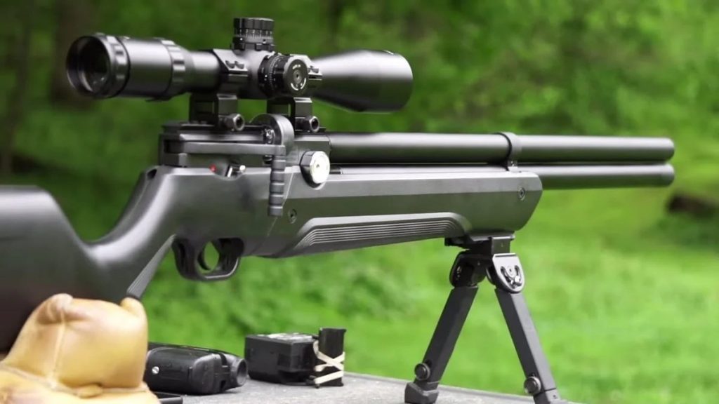 A black Air Venturi Avenger .25 Caliber air rifle with a scope and bipod is positioned on an outdoor table. It's surrounded by greenery, conveying a calm, focused atmosphere.