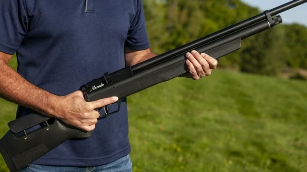 A person in a navy shirt holds a black Benjamin Marauder .22 Caliber air rifle outdoors on a sunny day. The background features a green field and trees, conveying a calm, rural setting.