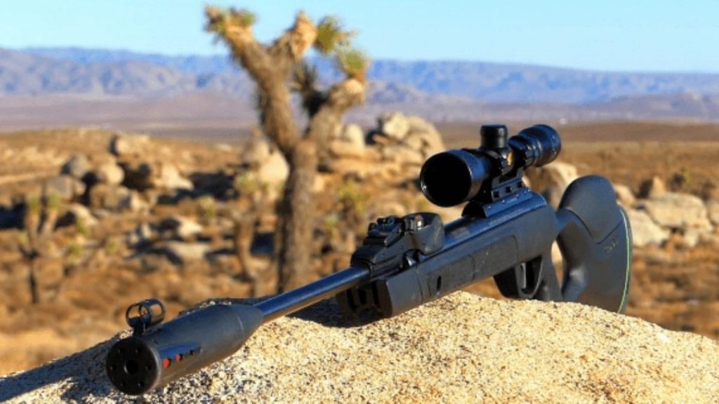 A black Gamo Swarm Magnum 10X GEN3i .22 Caliber hunting air rifle with a scope rests on a rock in a desert landscape, featuring sparse vegetation and distant mountains under a clear blue sky.