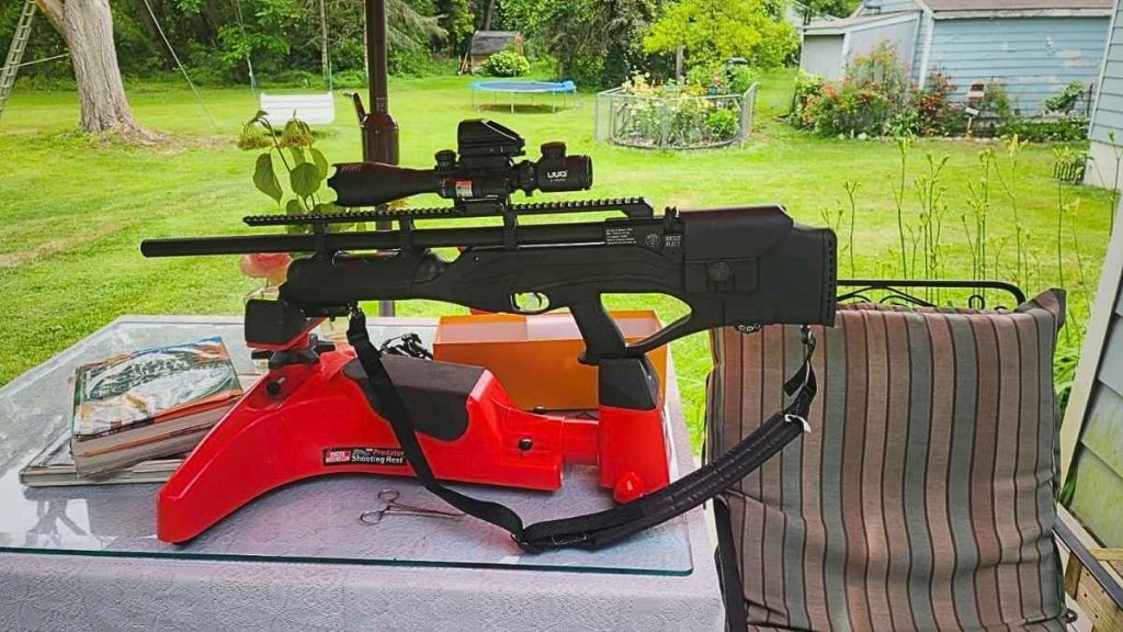 A black Hatsan Hercules Bully PCP .30 Caliber air rifle with a scope is resting on a red shooting stand atop a glass table on a porch. The background features a lush, green backyard.