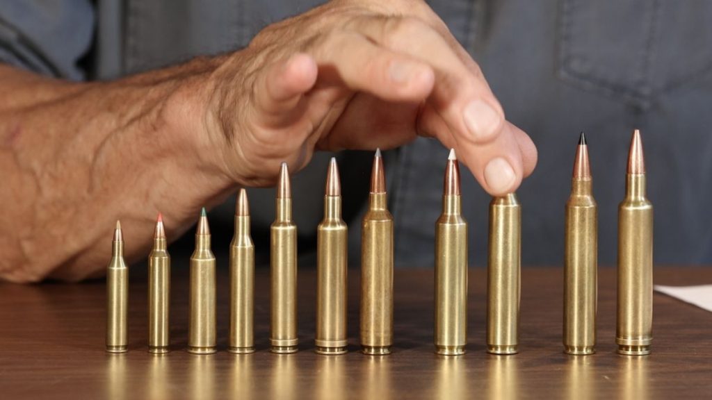 A variety of rifle cartridges arranged by size on a wooden surface. A hand is poised above them, with a focus on the different bullet tips.
