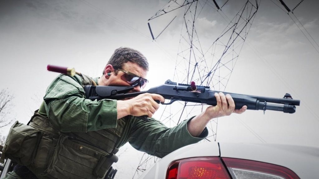 A tactical operator in green gear aims a Benelli M4 shotgun, standing next to a parked car with power lines in the background.