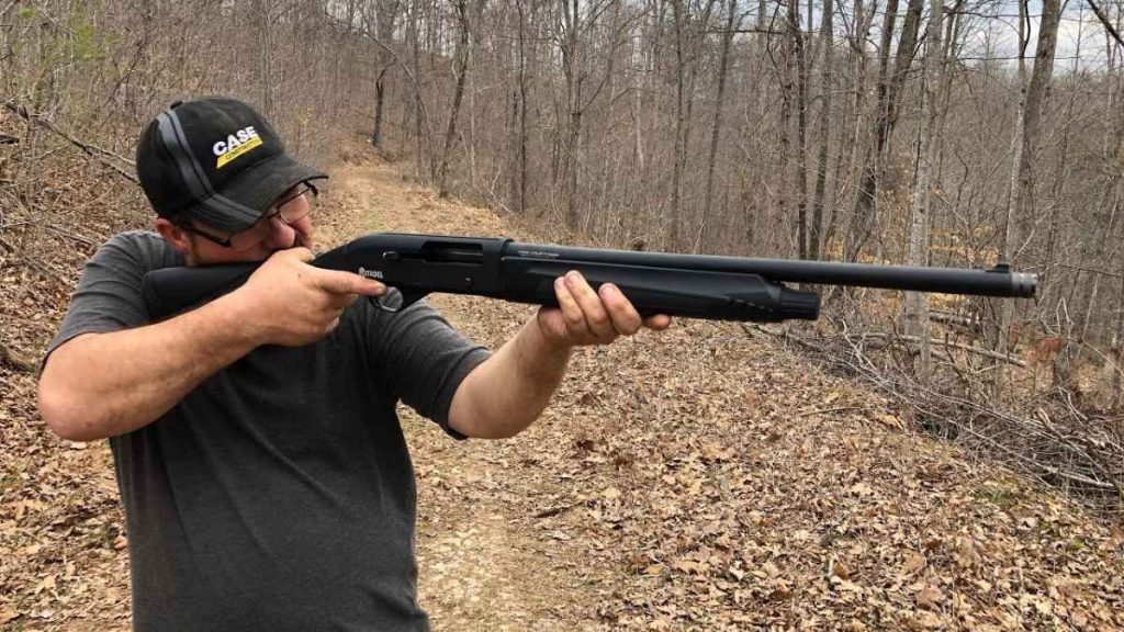 A person in a gray shirt and cap aims a Citadel Warthog 12-Gauge shotgun along a woodland trail, surrounded by bare trees and fallen leaves.