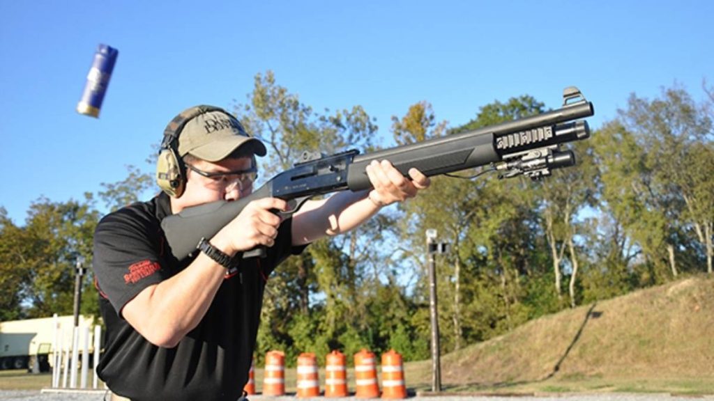 A person in safety gear aims a FN SLP Tactical shotgun outdoors, while a shot shell is ejected into the air against a clear blue sky.