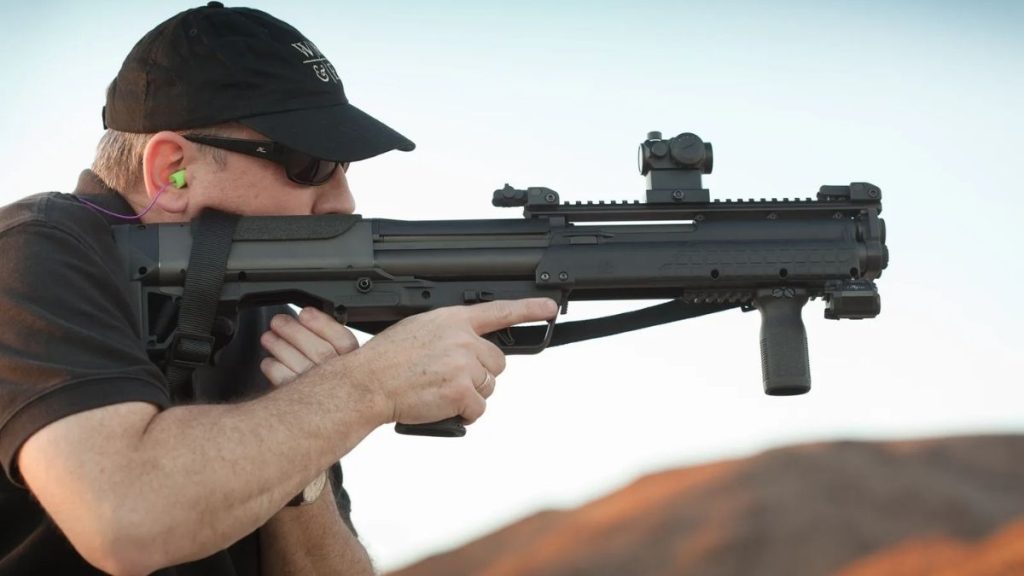 A person in a cap and ear protection aims a Kel-Tec KSG tactical shotgun at a shooting range, with a scenic backdrop of mountains.