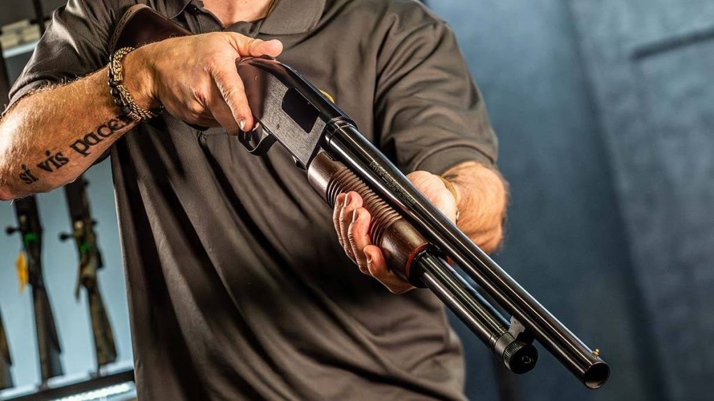 A person in a black shirt holds a Mossberg 500 shotgun, focusing on the weapon while surrounded by other firearms in the background.