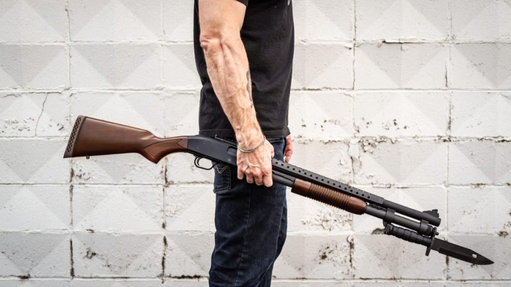 A person holding a Mossberg 590A1 shotgun with a wooden stock and a bayonet attachment, standing against a textured gray wall.