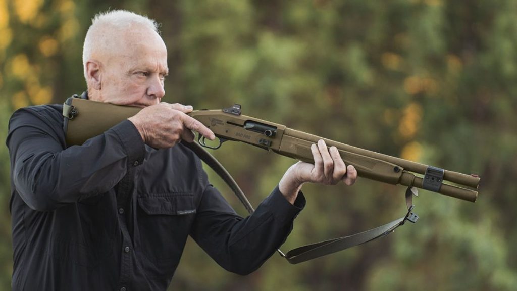 An older man in a black shirt aims a Mossberg 940 Pro Tactical shotgun outdoors, surrounded by blurred green foliage. The scene conveys focus and precision.