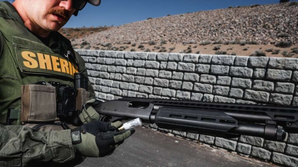 A sheriff's deputy in tactical gear examines a Remington 870 Tactical shotgun while holding a cartridge, with a stone wall and arid landscape in the background.