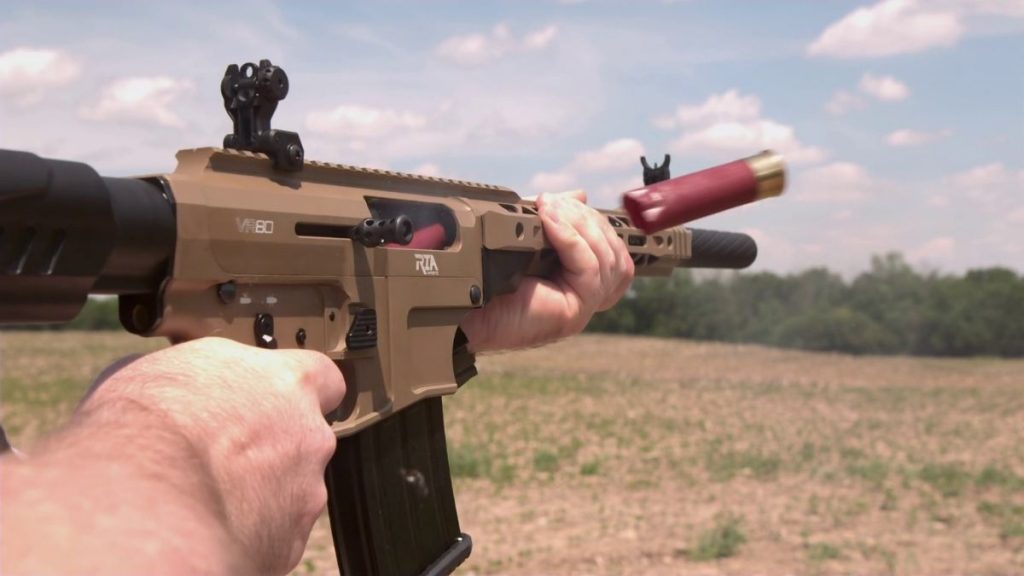 A person holds a tan Rock Island VR80 shotgun with a red shell chambered, aimed at a field under a blue sky with clouds.