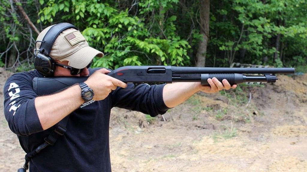 A person wearing tactical gear aims a Winchester SXP Defender shotgun outdoors, surrounded by greenery and gravel, demonstrating firearm handling.
