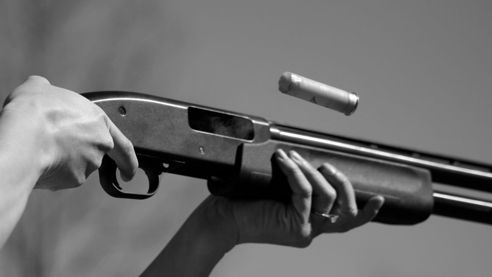The image shows a close-up view of a person's hands operating a best semi auto shotgun. The individual is pressing the trigger while their other hand supports the barrel. A shotgun shell is partially ejected from the chamber, capturing the moment just after firing. The photograph is in black and white, emphasizing the details and action of the shotgun mechanism.