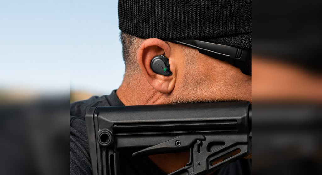 Close-up of a shooter wearing AXIL electronic ear plugs while aiming a rifle outdoors, with the earbud and logo clearly visible.