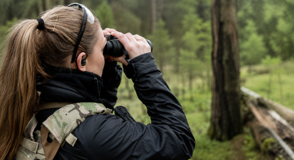 Woman wearing electronic ear plugs while using binoculars in a forest during a hunting or outdoor shooting trip.