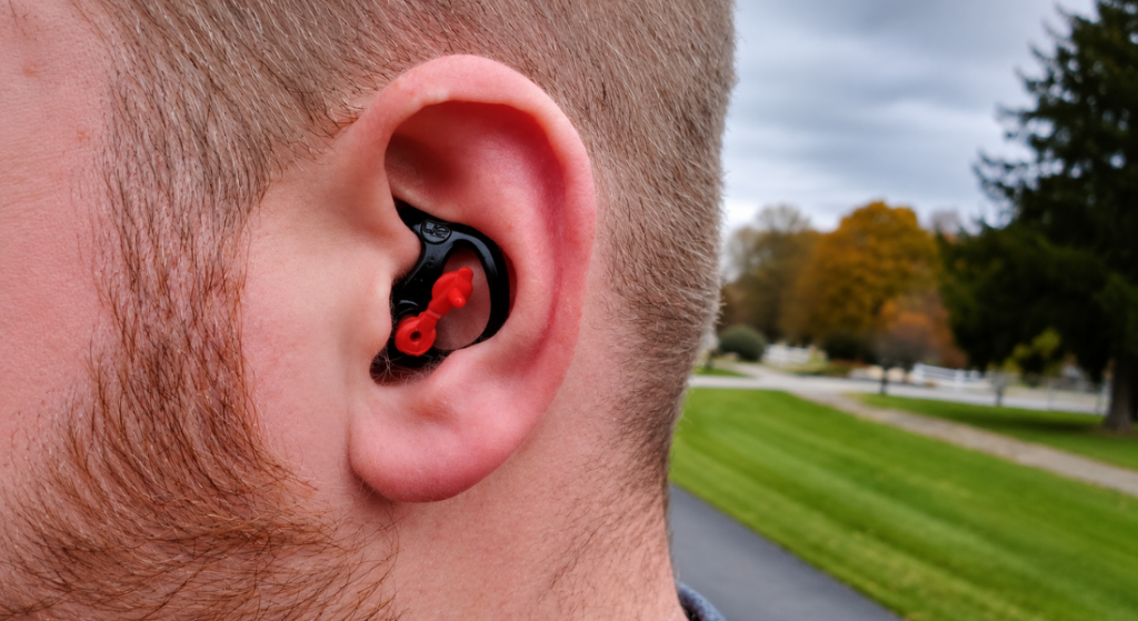 Close-up side view of a person wearing a black and red custom earplug, with the brand logo clearly visible, against a blurred outdoor background.