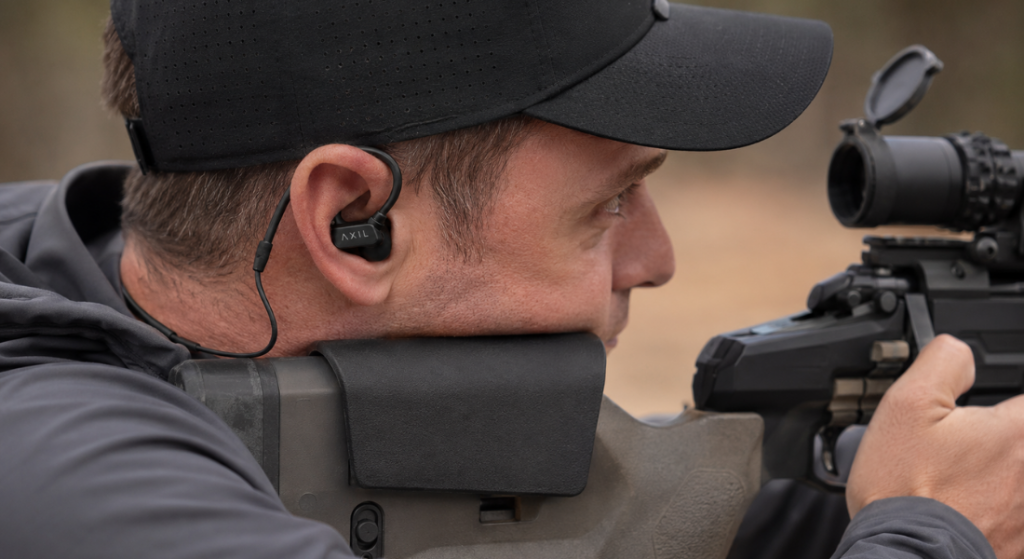 Man wearing AXIL electronic ear plugs while aiming a scoped rifle outdoors.