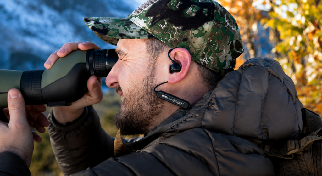 Hunter wearing ISOtunes Sport electronic ear plugs looks through a spotting scope in an outdoor hunting setting.