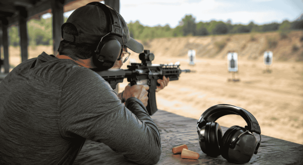 Shooter wearing earmuffs aiming a rifle at an outdoor range with extra hearing protection on the bench.