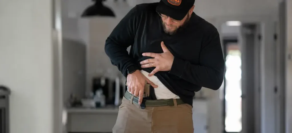 White man adjusting a Glock-style pistol in an appendix holster while wearing a black shirt and khaki pants indoors.