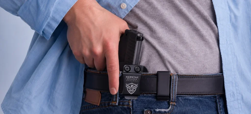 Close-up of a HAWKTAC Defense IWB holster clipped to a belt, with a concealed handgun at the waistband under a blue shirt.