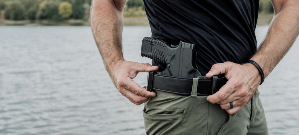 Man adjusting an OWB holster with a compact pistol secured on his belt near a lake.