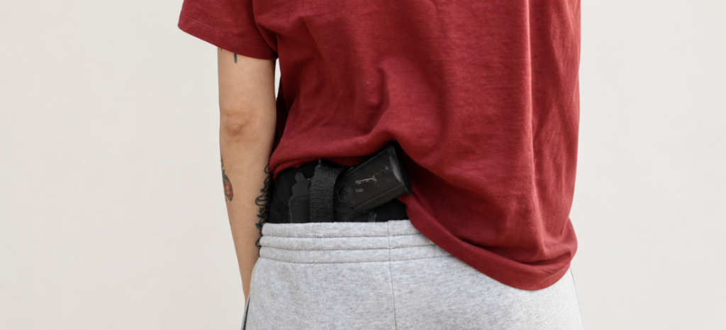 Person wearing a red T-shirt and gray joggers with a black concealed carry holster and handgun tucked at the waistband.