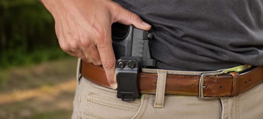 Close-up of a person carrying a black waistband holster with a handgun, wearing a dark shirt, tan pants, and a brown belt.