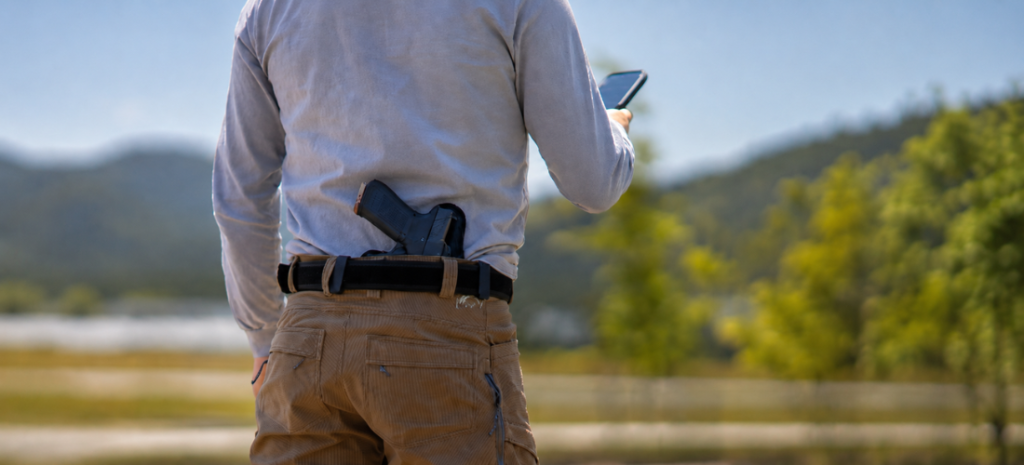 Man carrying a concealed S&W Shield-style pistol in a waist holster while standing outdoors with a phone in hand.
