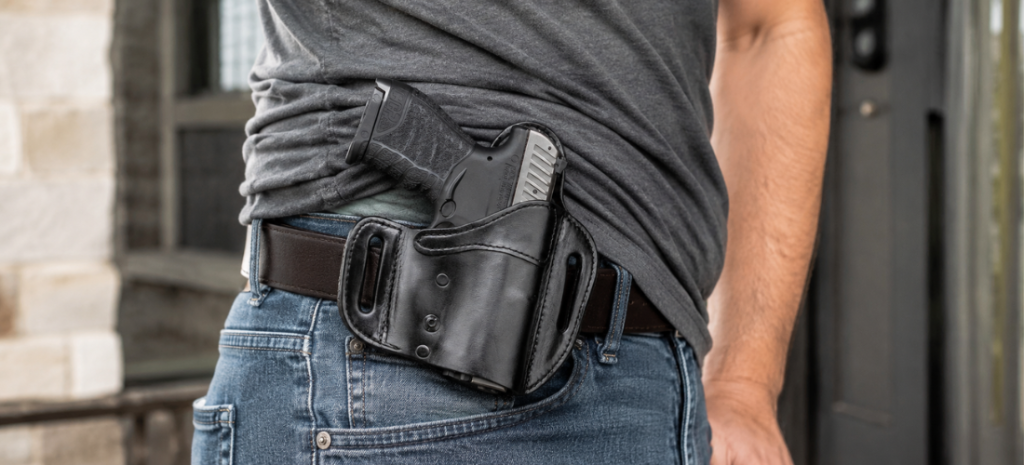 Man wearing an OWB leather holster with a compact pistol visible on a belt under a lifted gray shirt.