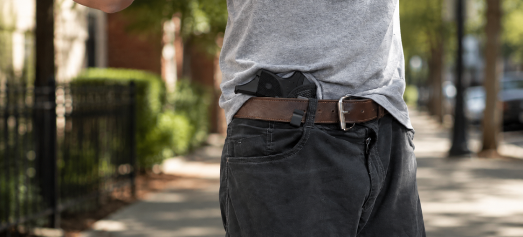 Man walking outdoors with a compact pistol visible in an IWB holster under a gray shirt.