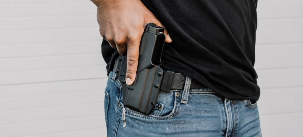Close-up of a concealed carry holster with a handgun worn at the waistband under a black shirt and blue jeans.