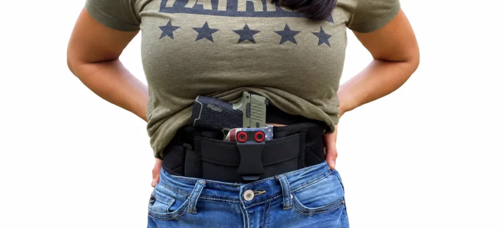 Woman wearing a black waistband holster with a handgun and magazine under a lifted green T-shirt.