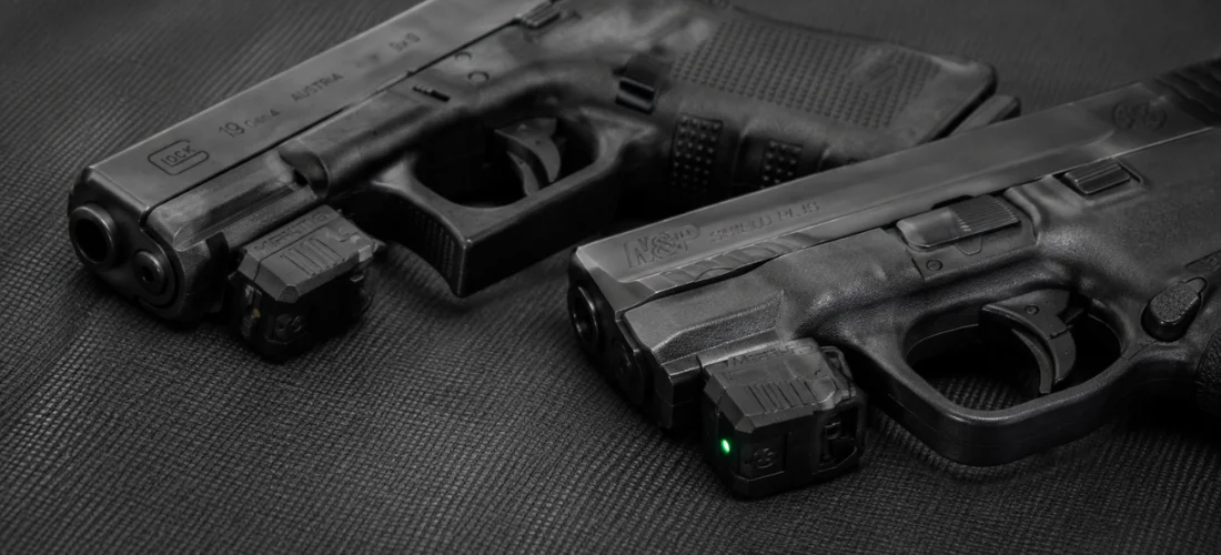 Mantis X3 and X10 Elite training sensors mounted beneath two unloaded pistols on a dark shooting mat.