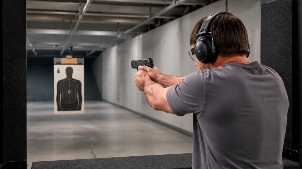 Man wearing shooting earmuffs and safety glasses while aiming a handgun at a dummy target in an indoor range.