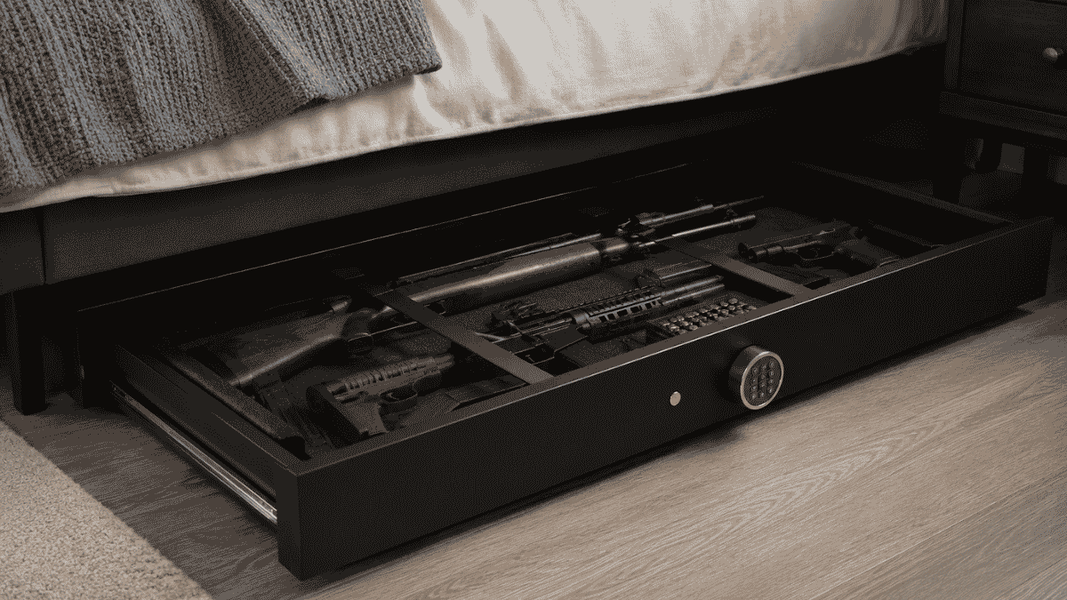 Open under-bed gun safe with rifles, handguns, ammunition, and accessories stored in a bedroom.