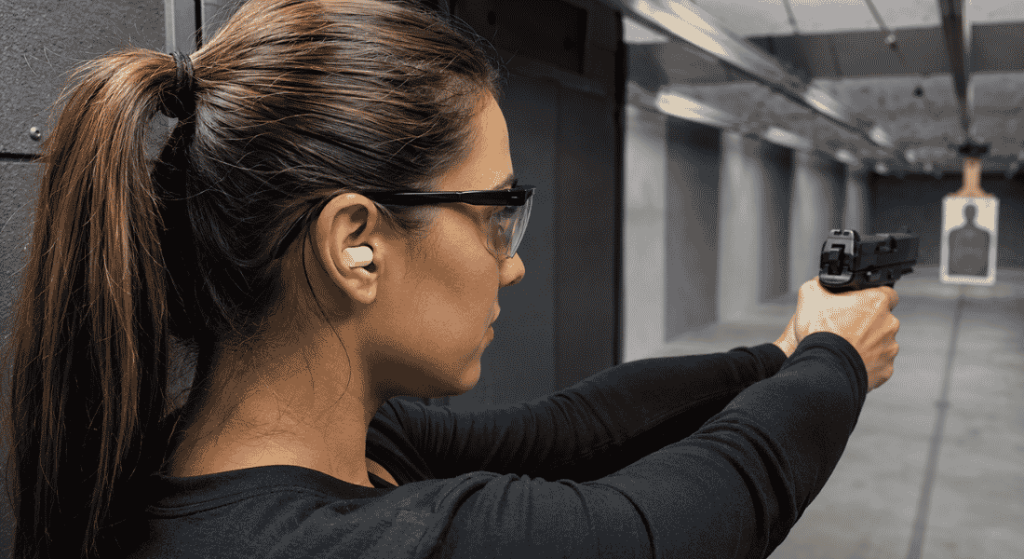 Woman wearing shooting earmuffs and safety glasses while aiming a handgun at an indoor shooting range target.