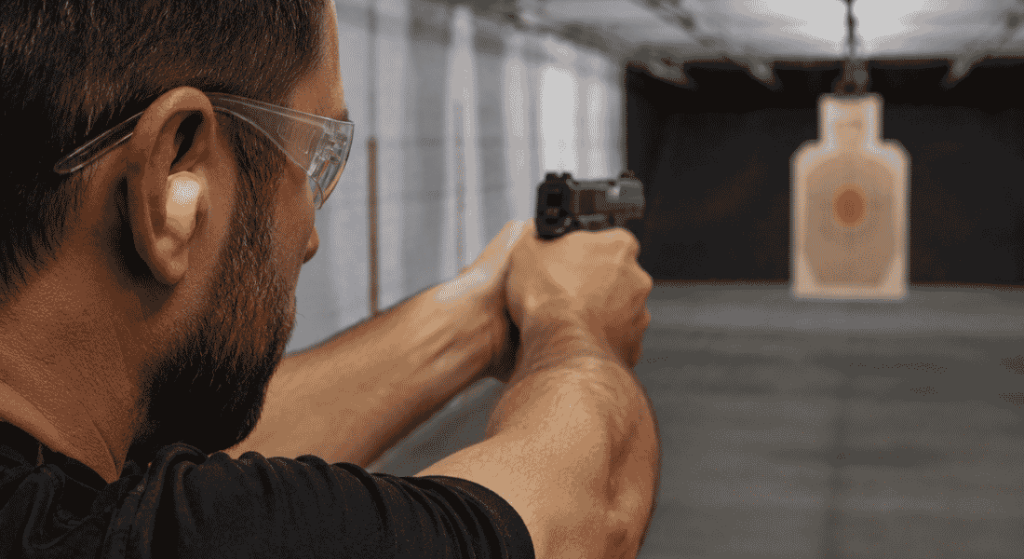 Man wearing foam earplugs and safety glasses while aiming a handgun at a dummy target in an indoor shooting range.