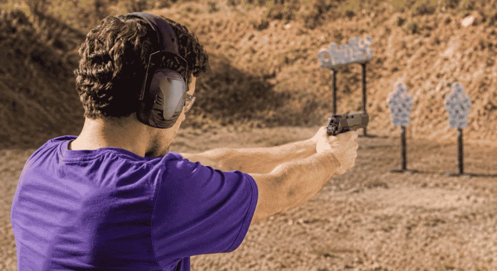 Man wearing shooting earmuffs and safety glasses while aiming a handgun at outdoor range targets.