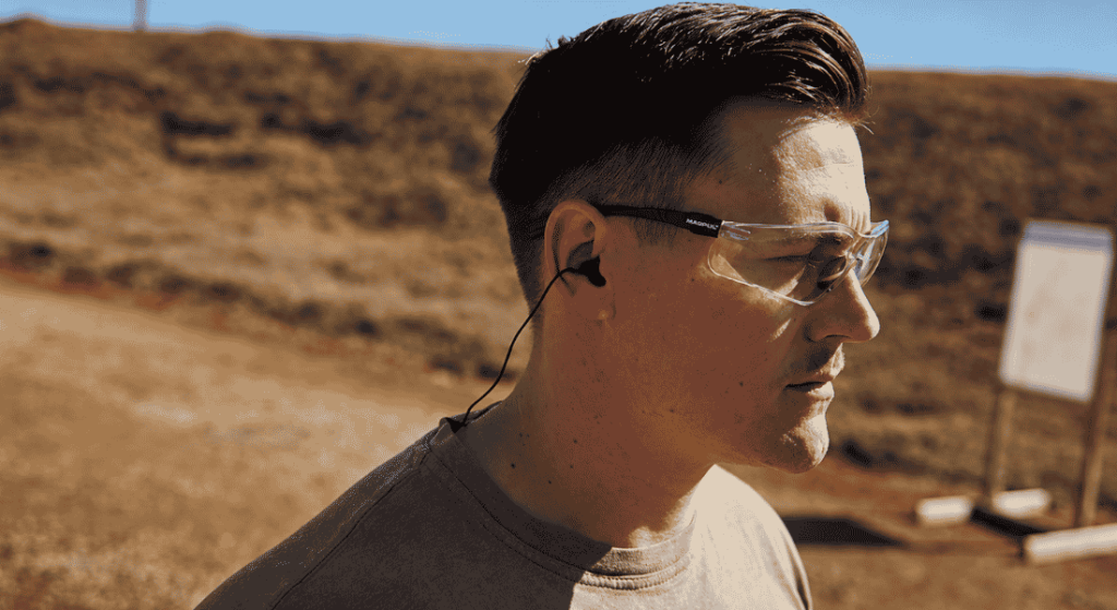 Man wearing clear safety glasses and black earplugs at an outdoor shooting range.