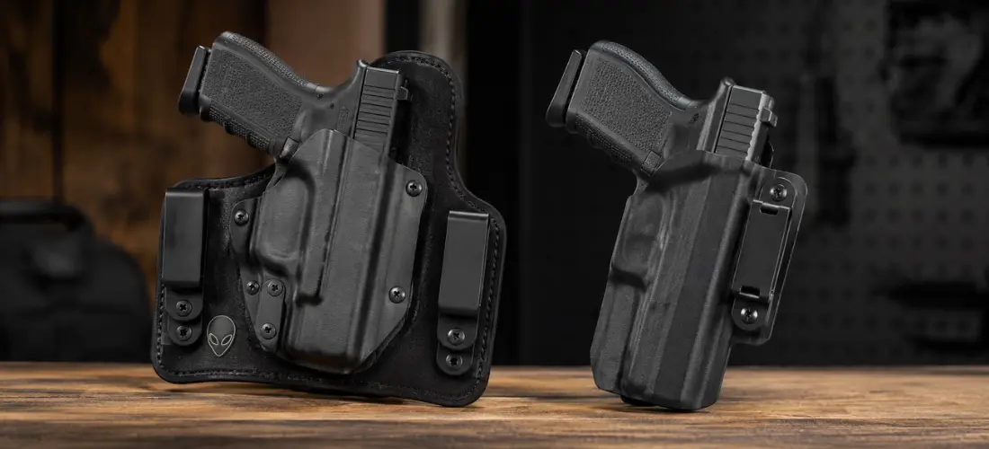 Side-by-side comparison of a hybrid concealed-carry holster and a slim Kydex holster with handguns on a wooden surface against a dark workshop background.
