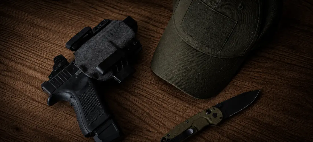 Glock 19-style pistol in an AIWB holster with a tactical cap and folding knife on a dark wooden tabletop.