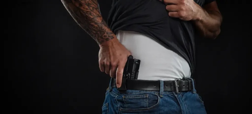 Tattooed man carrying a Glock-style pistol in a black appendix holster with jeans and a black shirt.