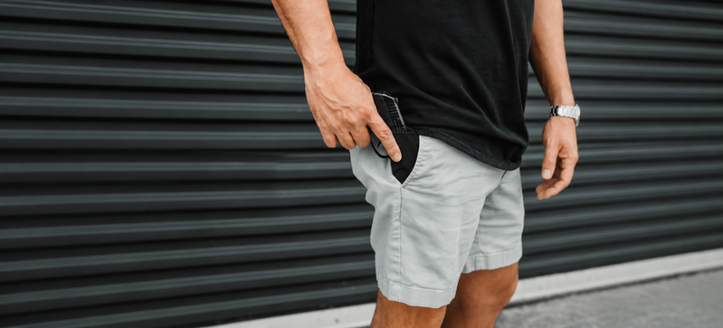 Man placing a compact pistol in a black pocket holster inside the front pocket of light gray shorts.