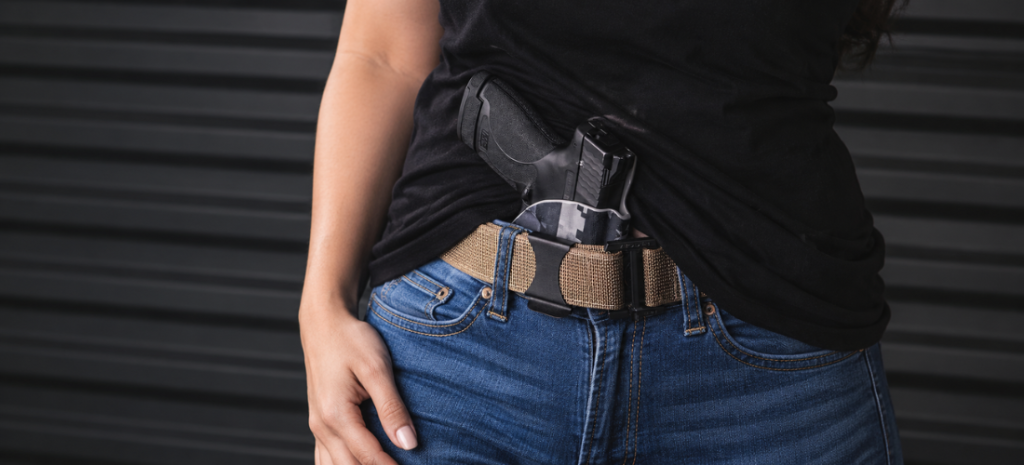 Woman wearing a black shirt carrying a compact pistol in a visible IWB holster on a tan belt with blue jeans.
