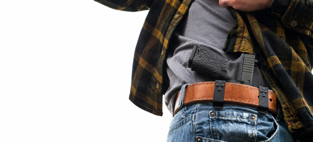 Close-up of a person carrying a compact handgun in an appendix holster with a brown leather belt, plaid flannel shirt, and blue jeans.